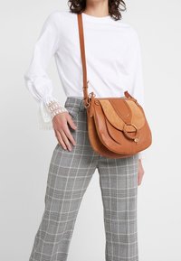 Brun læder- og ruskinds crossbody taske med en cirkulær hardwaredetalje, flad bund og justerbar skulderrem, båret med ternet wide-leg bukser.