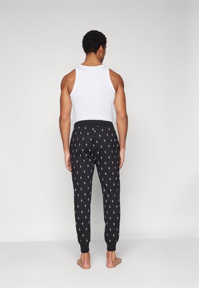 Pantalons de pyjama noirs avec un motif de logo blanc en répétition, comportant une taille élastique et des poignets, portés avec un débardeur blanc.