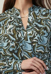 Blusa fluida in blu, verde e beige con motivi a vortice; tessuto con una consistenza morbida; presenta un colletto alto e polsini con bottoni.