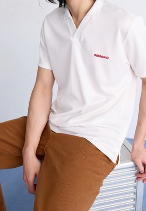Homme portant un polo blanc Adidas avec un logo rouge, pantalon marron, assis sur un banc en métal argenté.