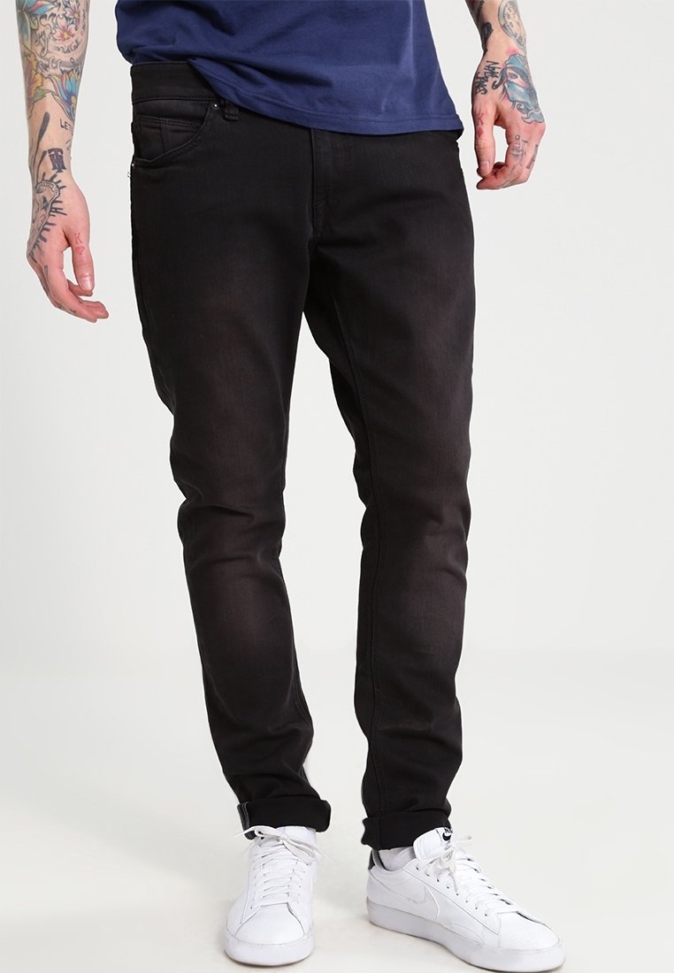 Homme portant un jean slim noir, des baskets blanches et un t-shirt bleu, avec des bras tatoués visibles sur un fond clair uni.