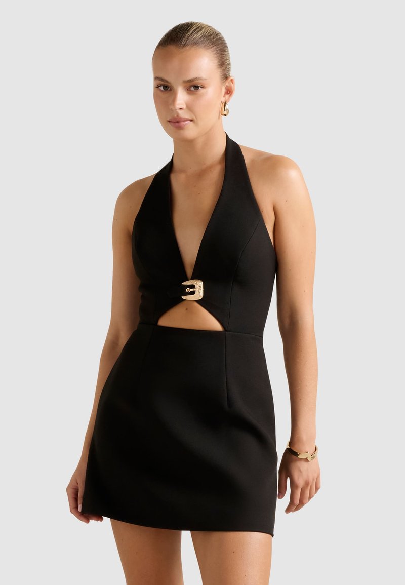 Donna che indossa un mini abito nero con scollo all'americana, fibbia dorata e dettagli cut-out in vita, orecchini dorati e un braccialetto dorato.