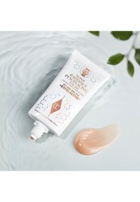 Tube de crème de resurfaçage facial étiqueté "Super Radiance Resurfacing Facial" avec une trace de produit crémeux sur une surface d'eau ondulée et des feuilles vertes à proximité.