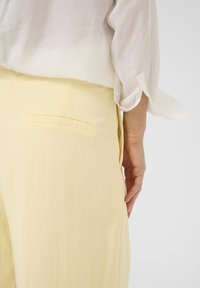 Pantaloni giallo chiaro con una texture liscia, dotati di una tasca posteriore e di un design a gamba larga, abbinati a una blouse bianca semi-trasparente.