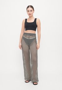 Top corto nero abbinato a pantaloni a palazzo ampi in tessuto trasparente con motivo zigzag bianco e nero. La modella è in piedi su uno sfondo semplice, indossando sandali neri semplici.