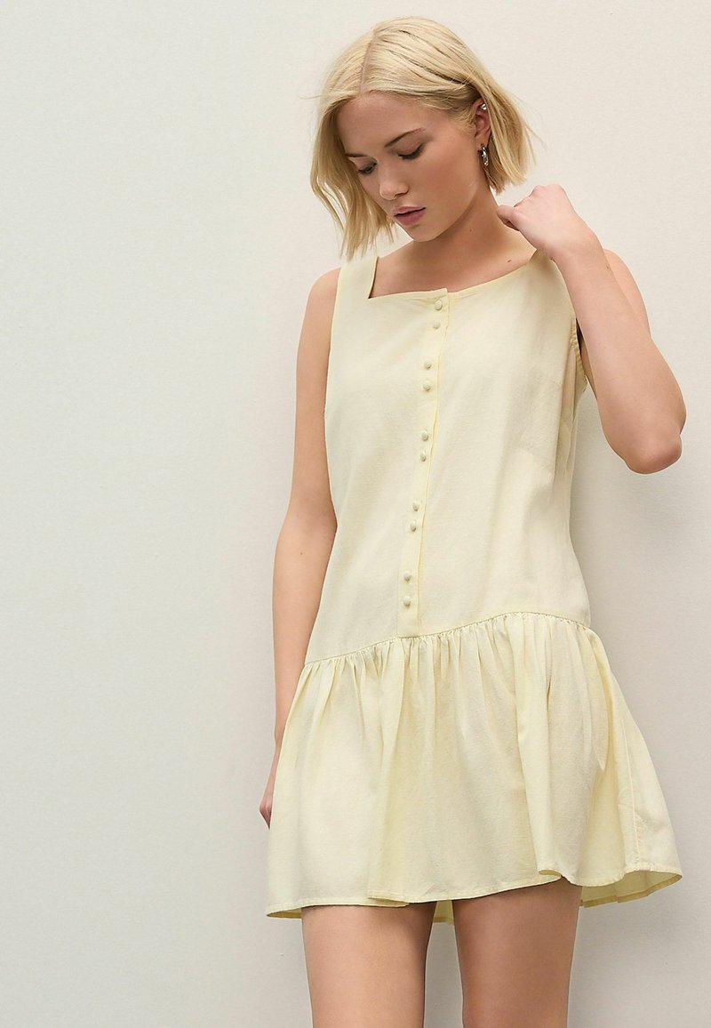 Robe sans manches jaune avec un décolleté carré, des boutons sur le devant et une jupe évasée à volants. Fabriquée en tissu léger et doux.