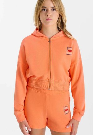 Young woman wearing an orange zip-up hoodie and matching shorts with small stamp-like patches on chest and thigh.