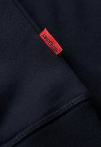 Tessuto felpa blu navy con una texture liscia, dotato di un'etichetta rossa "Superdry". Dettagli di cucitura visibili lungo i bordi.
