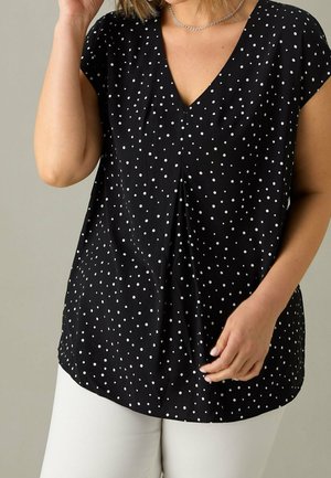 Kobieta ubrana w czarną bluzkę z krótkim rękawem w białe kropki i białe spodnie, pozująca z jedną ręką przy twarzy, a drugą przy talii.