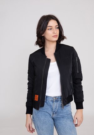 Giacca bomber nera con chiusura a zip, polsini a coste e un'etichetta arancione sul lato sinistro. Presenta una tasca laterale e una texture liscia.