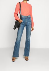 Persona che indossa una blusa a maniche lunghe testurizzata color corallo, jeans flared blu a vita alta, tacchi a strappo neri e porta una borsa a tracolla rotonda nera.