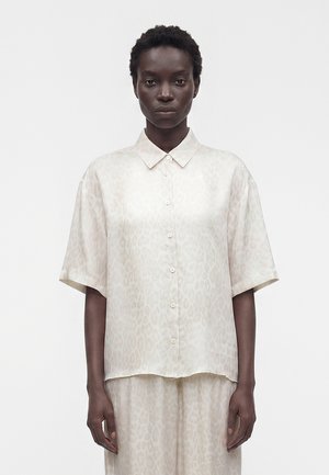 Personne à la peau foncée avec des cheveux courts portant une chemise ample en satin beige clair à boutons avec un subtil imprimé léopard et un pantalon assorti.