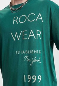 Grøn bomulds T-shirt med hvid tekst: "ROCA WEAR ESTABLISHED New York 1999," med en glat tekstur og korte ærmer. Sølv kæde halskæde synligt.