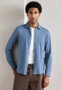 Camisa azul-claro de botão com colarinho e mangas compridas, combinada com uma t-shirt branca, calças castanho-escuras e um cinto preto.