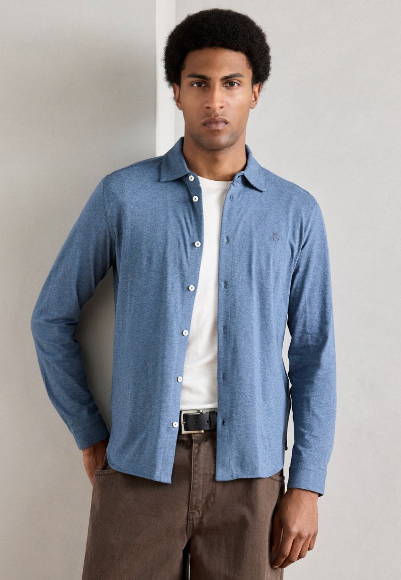 Camisa azul-claro de botão com colarinho e mangas compridas, combinada com uma t-shirt branca, calças castanho-escuras e um cinto preto.