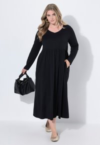 Robe noire à manches longues avec un décolleté en V, taille froncée et poches latérales. Associée à un sac à main noir et des chaussures slip-on beiges.