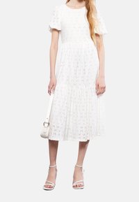 Kobieta w białej, teksturowanej sukience midi z bufkami, trzymająca białą torebkę i nosząca białe sandały na obcasie z paskami.