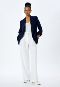 Blazer navy con spalle strutturate, abbinato a pantaloni bianchi a vita alta. Il blazer presenta maniche con bottoni; tessuto testurizzato su tutta la superficie.