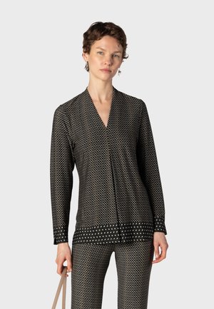 Femme aux cheveux courts et bouclés portant un haut noir à manches longues à motif et un pantalon assorti, tenant une sangle de sac beige sur un fond uni.