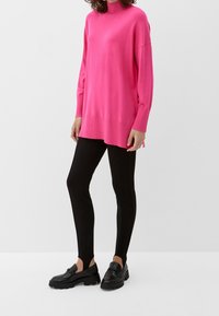 Fuchsia übergroßer Pullover mit hohem Kragen, langen Ärmeln und gerippten Details, kombiniert mit schwarzen eng anliegenden Hosen und schwarzen Loafers.