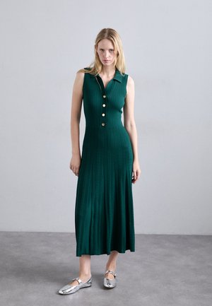 Mujer de pie con un vestido de punto verde sin mangas, botones dorados y zapatos planos plateados, sobre un suelo gris y un fondo de pared gris lisa.