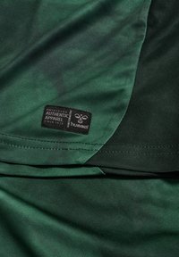 Sportowa odzież z ciemnozielonej tkaniny o gładkiej fakturze, z czarną metką "hummel" i widocznymi detalami szwów.