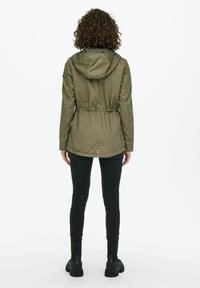 Olivgrön vattentät jacka med huva, dragsko i midjan och rundad nederkant. Bärs över svarta skinnyjeans och stövlar.