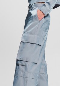 Svijetloplave, sjajne cargo hlače s više džepova i šivenim detaljima. Imaju crnu naljepnicu "KARL LAGERFELD JEANS" blizu rubova.