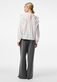 Blouse en coton blanc avec détails à volants sur les épaules et dos en keyhole, associée à un pantalon gris à jambe large. Texture lisse et coupe décontractée.