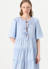 Lys blå lagdelt kjole med korte ermer, teksturert stoff, og en nøkkelhullfront knyttet med to sløyfer. Distinkt bølgete mønster gjennom hele.