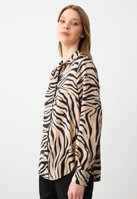 Lange mouw blouse in beige satijn met zwarte zebrapatronen, met een knoopsluiting aan de voorkant en een relaxte pasvorm. Soepele textuur en kraagdetail.