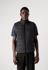 Black padded vest with a high collar, featuring a zip closure and subtle logo detail, worn over a dark short-sleeve shirt.
