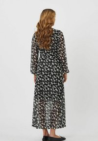 Robe longue et légère présentant un motif abstrait noir et blanc. Elle a des manches longues et une silhouette fluide, atteignant le milieu du mollet. Chaussures plates noires.