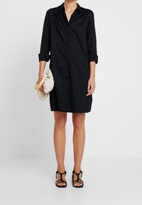 Robe chemise noire à manches longues, fermeture à boutons sur le devant et fentes sur les côtés. Associée à un sac blanc texturé et des sandales noires à lanières.