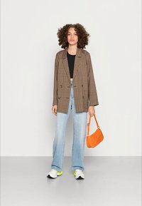 Jovem mulher com cabelos encaracolados, vestindo um blazer castanho xadrez, top cropped preto, jeans azul claros e tênis brancos, segurando uma bolsa laranja.