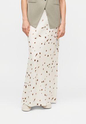 Femme portant une jupe longue blanche à taches marron, un gilet beige et des baskets blanches, debout devant un fond clair uni.