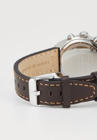 Bracelet de montre en cuir marron avec des coutures beige, boucle en acier inoxydable et gravure. Une texture lisse et des trous rectangulaires pour l'ajustement.