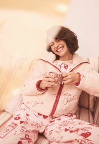 Kremna flis jakna z rdečimi poudarki in vezenim napisom, združena z rožnatimi pajkicami z ročno risanimi vzorci srčkov. Drži rožnato skodelico.