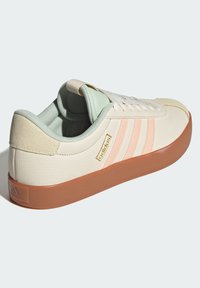 Vita lädersneaker med ljusgrön inredning, vita snören, rosa ränder och en brun gummisul. Guldtryck på sidan.