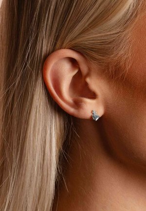 Silberner Ohrstecker in Herzform mit kleinem klaren Akzent, an einem hellhäutigen Ohr getragen. Glatte Oberfläche und minimalistisches Design.