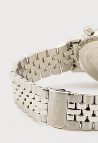 Pulseira de relógio Timex em metal prateado com fecho, apresentando segmentos interligados e parte da caixa do relógio com a coroa visível.