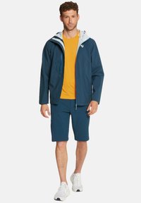 Marinblå jacka med ljusgrå huva, orange t-shirt och marinblå shorts. Tyget är smidigt; har dragkedjor och fickor. Vita sneakers.