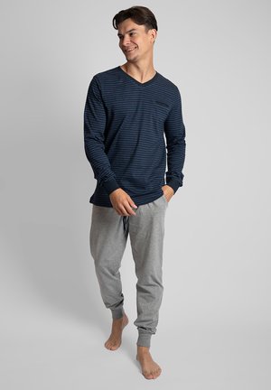 Mannen navy gestreepte langwerpige V-hals shirt gecombineerd met grijze joggingbroeken, met een trekkoord in de taille en enkelmanchetten. Blootsvoets.