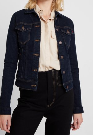 Kvinde iført en mørkeblå denimjakke over en beige skjorte med knapper og sorte bukser med synlige gule sømme.