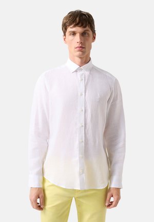 Man facing forward wearing a white long-sleeve button-up shirt and light yellow pants against a plain background.