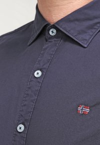 Mörkblå skjorta med knappar, utrustad med klassisk krage, ljusblåa knappar och en liten broderad norsk flagga på bröstet.