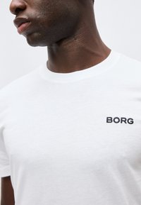 Κοντινό πλάνο ενός ατόμου που φοράει λευκό μπλουζάκι με το κέντημα "BORG" μαύρο στο αριστερό στήθος.