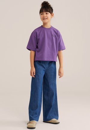 Enfant aux cheveux foncés attachés, portant un haut violet à manches courtes, un jean bleu à jambes larges et des chaussures beige à enfiler, debout et souriant.