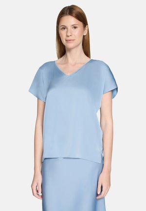 Junge Frau mit glatten, hellbraunen Haaren, die eine hellblaue Kurzarm-Bluse mit V-Ausschnitt und einen passenden Rock trägt, vor einem einfarbigen Hintergrund stehend.