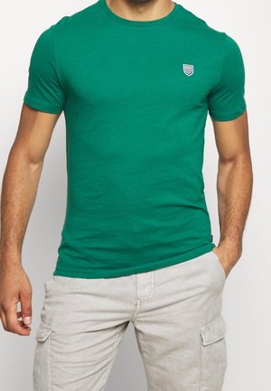 Mann trägt ein eng anliegendes grünes Kurzarm-T-Shirt mit kleinem Brustlogo und hellbeige Cargo-Hose, eine Hand in der Tasche.